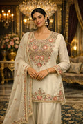 Woman in a cream embroidered Anarkali suit with floral motifs, wearing statement earrings, standing in a luxurious room.