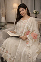 Woman in cream embroidered ethnic suit with floral dupatta sitting on sofa reading a book