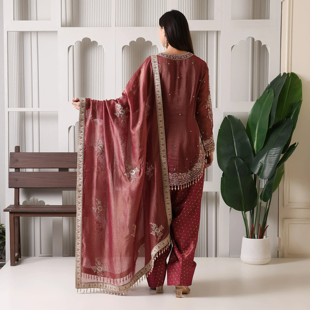 Woman in maroon embroidered ethnic suit with dupatta, standing indoors near potted plant.