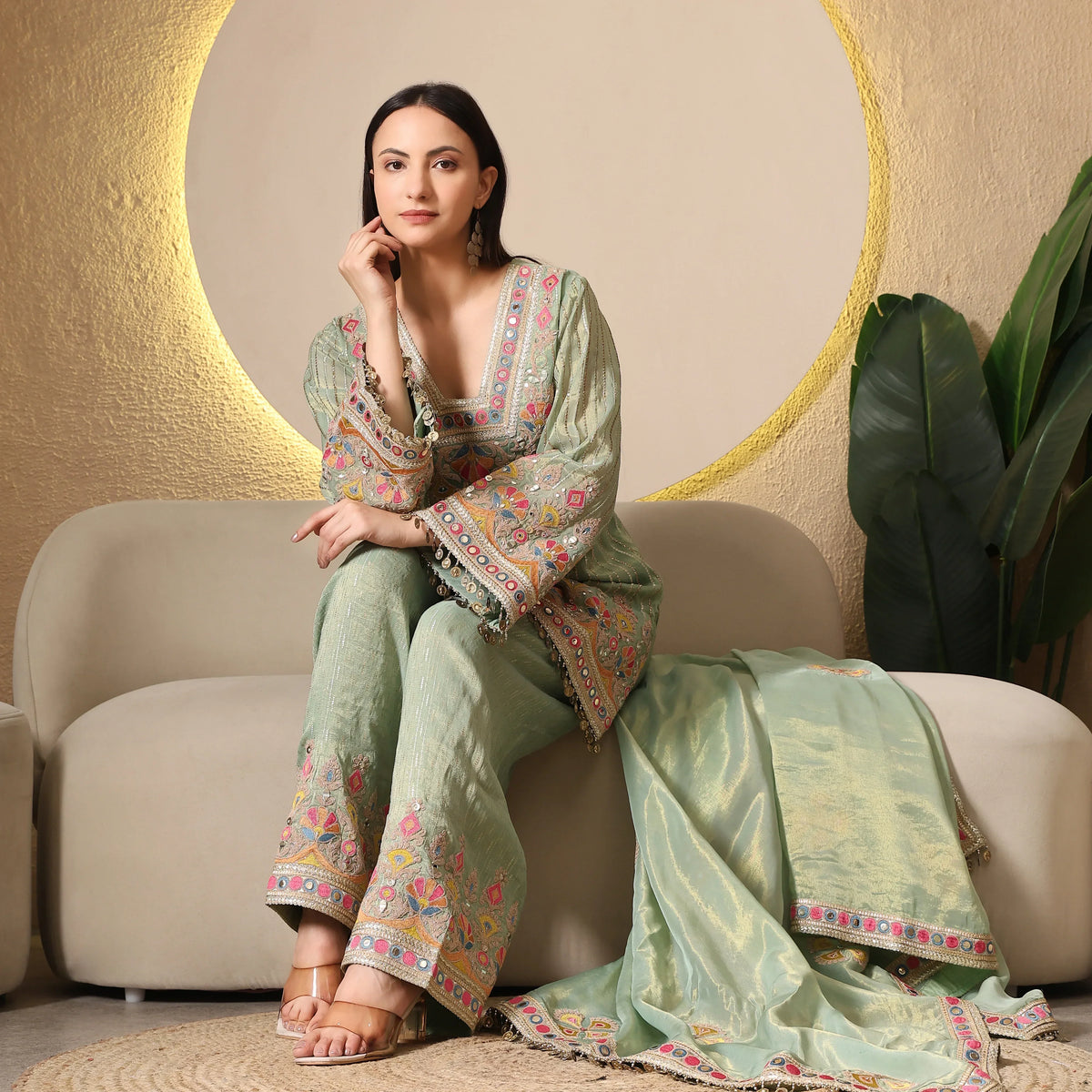 Woman in mint green embroidered ethnic suit with dupatta, sitting on beige sofa.