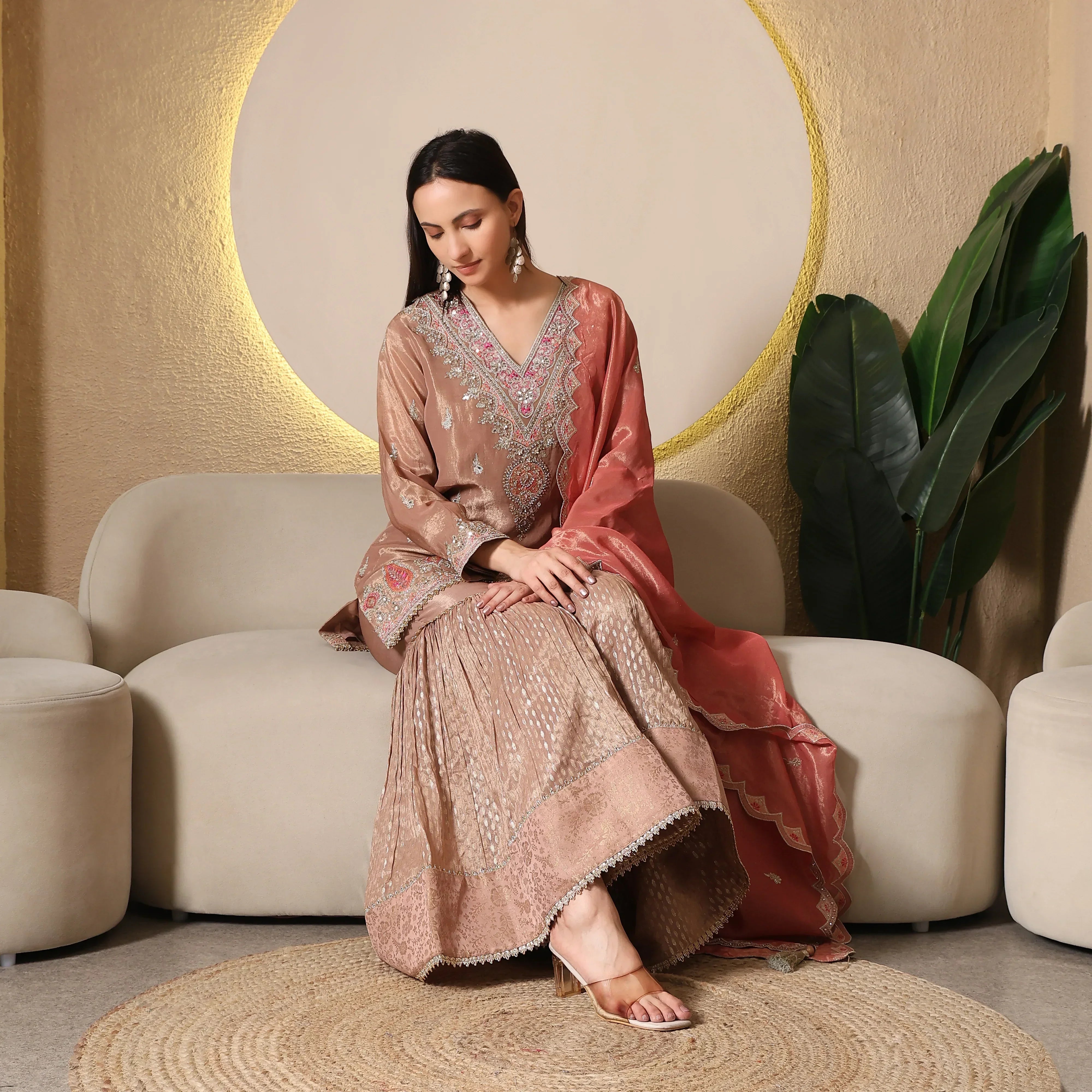 Woman wearing embroidered Banarasi Anarkali suit with dupatta sitting on beige sofa