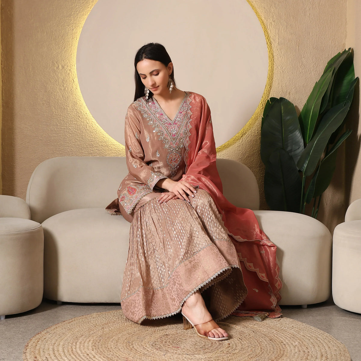 Woman wearing embroidered Banarasi Anarkali suit with dupatta sitting on beige sofa