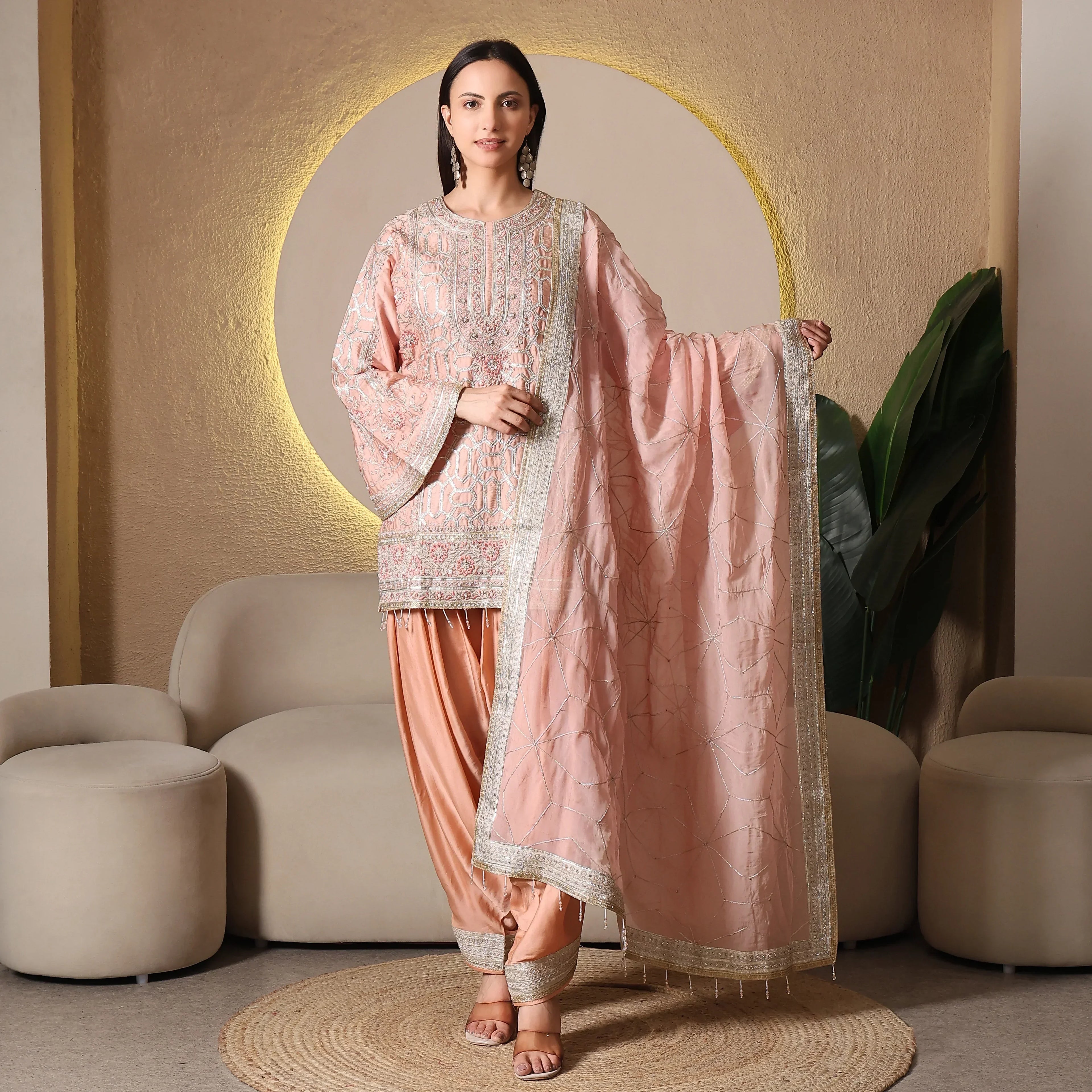 Woman in blush pink embroidered ethnic suit with matching dupatta, festive indoor setting.