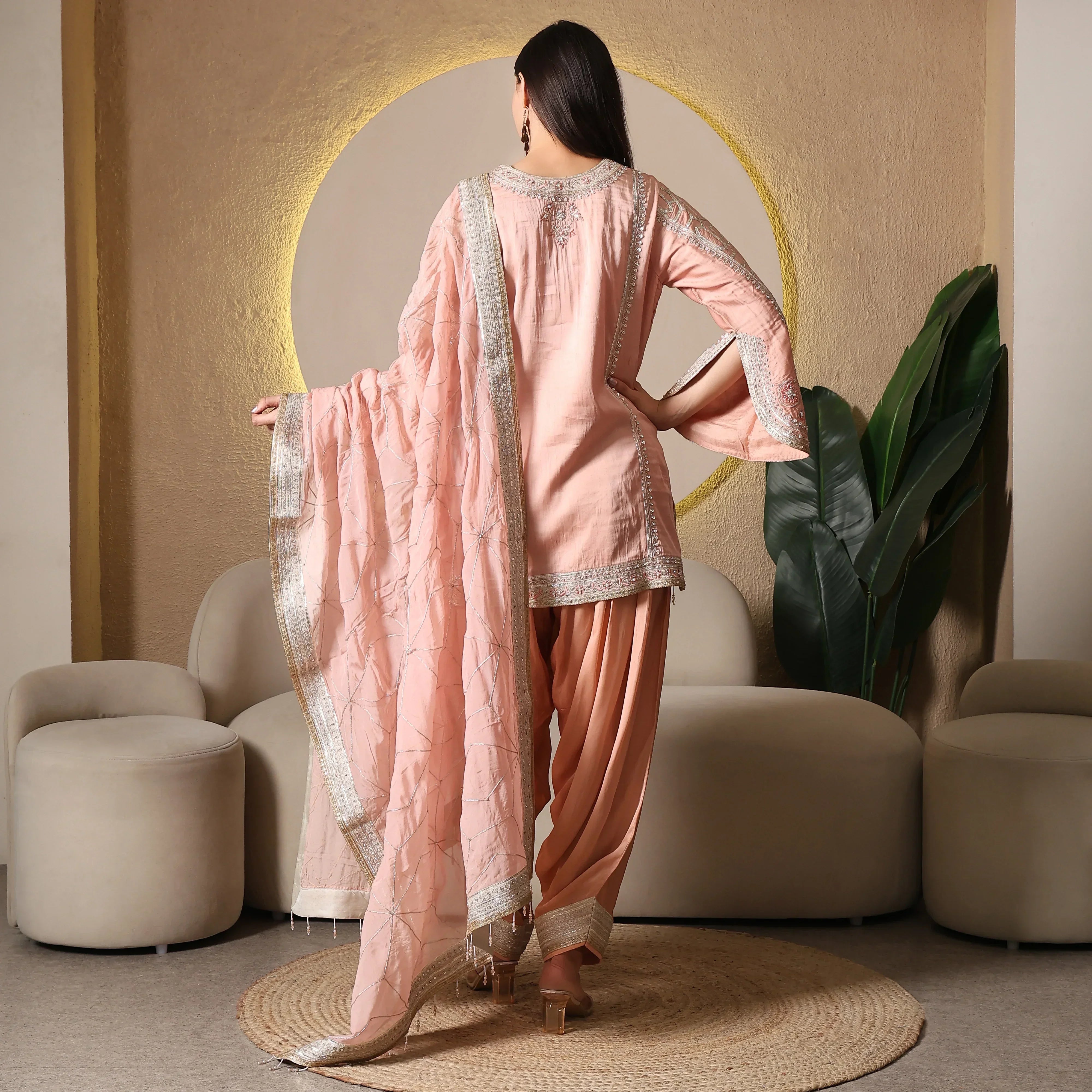 Woman in pastel pink ethnic suit with embroidered dupatta, standing in modern lounge