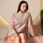 Woman wearing pastel peach embroidered ethnic suit, sitting on a beige sofa indoors.