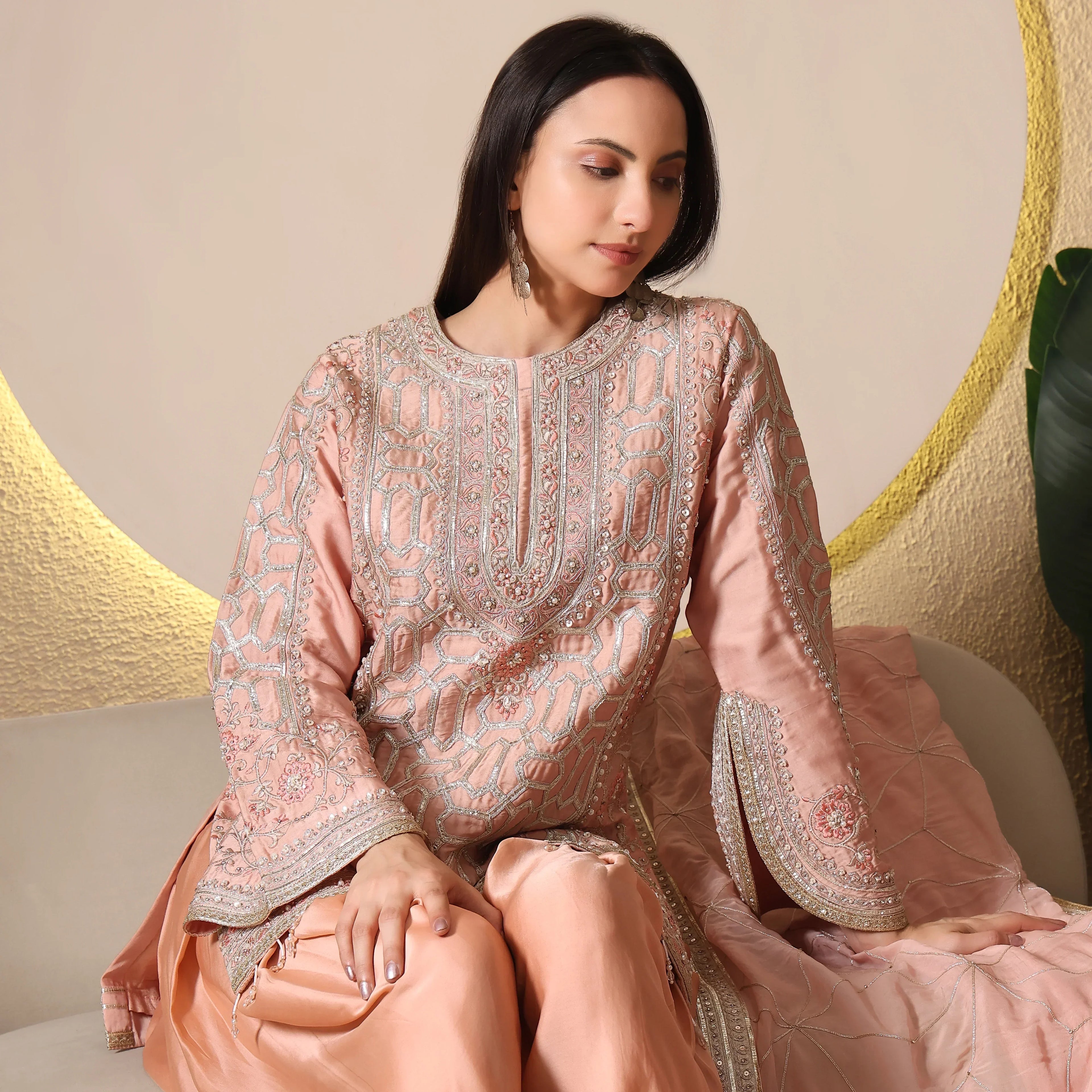 Woman wearing pastel peach embroidered ethnic suit, sitting on a beige sofa indoors.