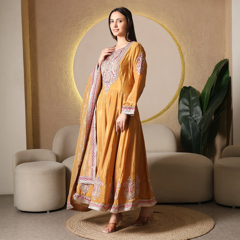 Woman wearing a mustard yellow embroidered Anarkali suit with dupatta, standing in a modern living room.