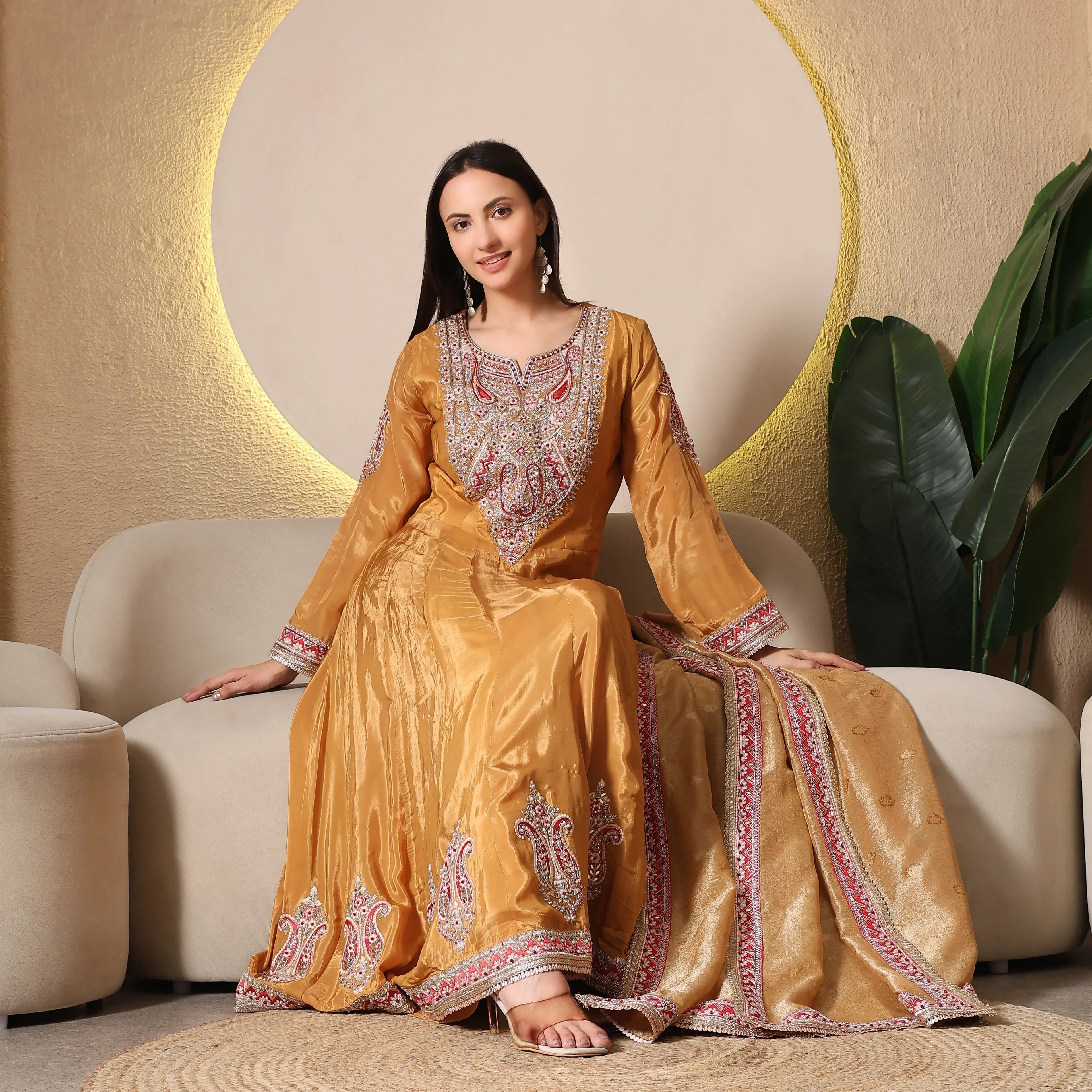 Woman wearing gold embroidered Anarkali suit with dupatta, sitting on a beige sofa