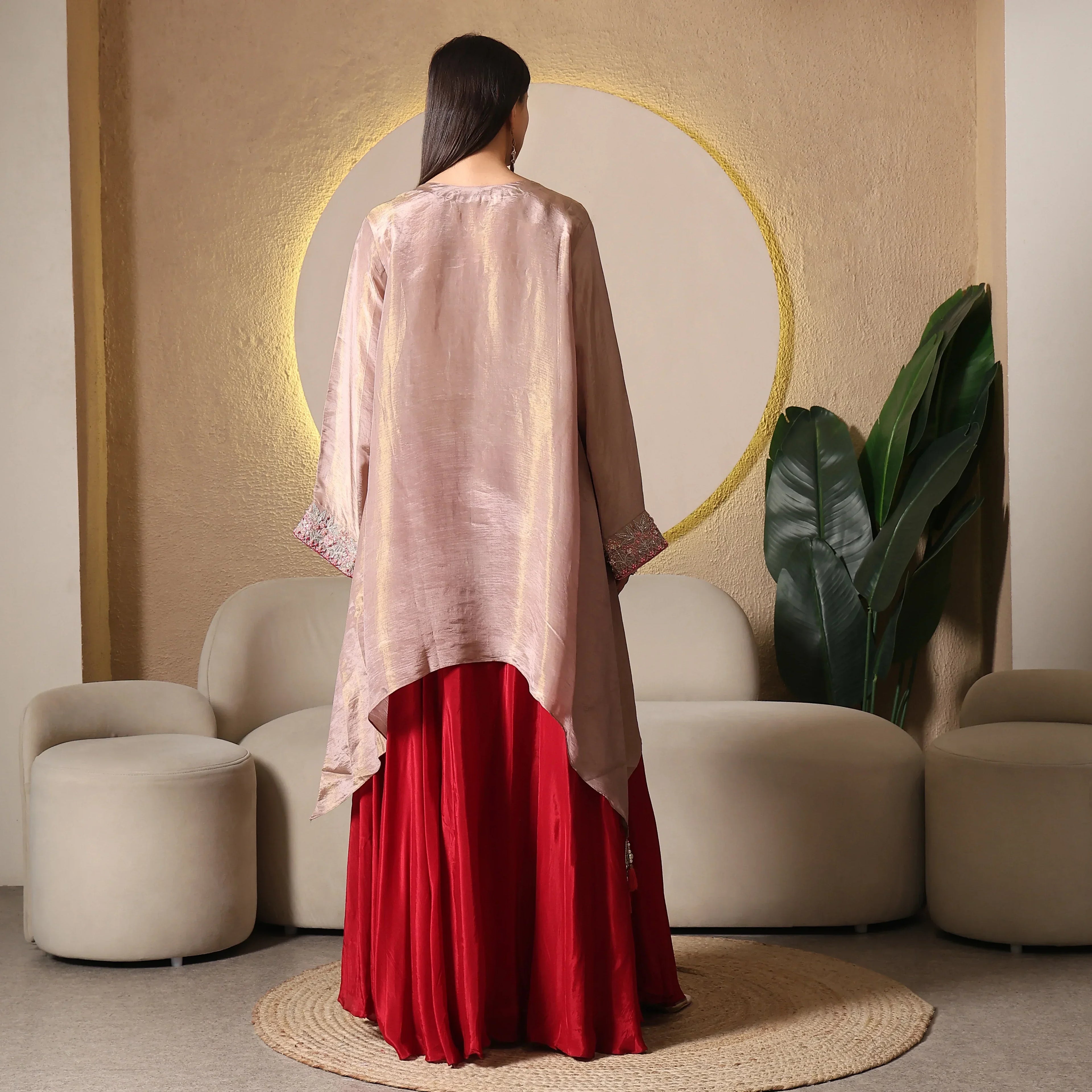 Woman in a flowy red Anarkali suit with a gold-tone high-low jacket, standing in a modern ethnic living room