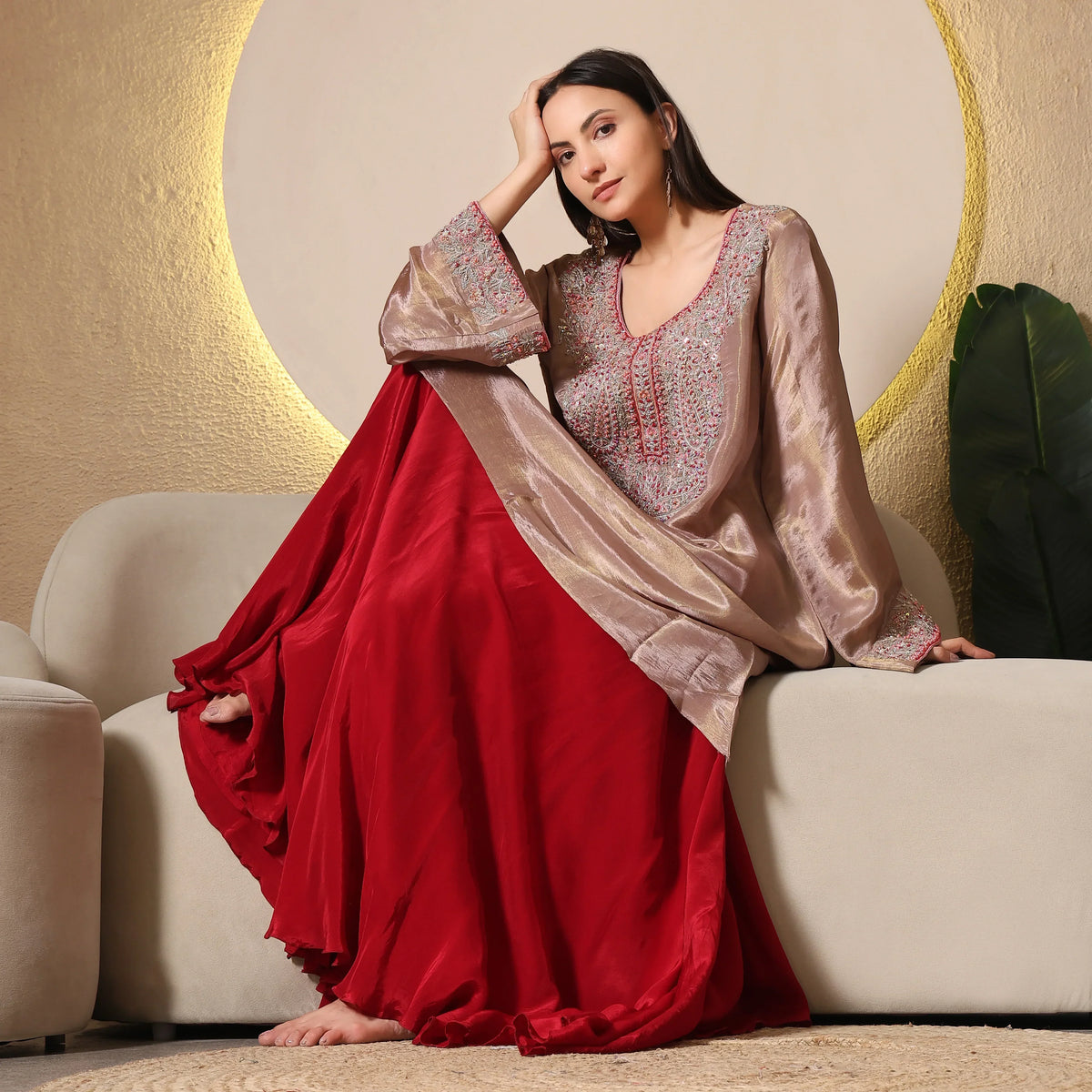 Woman in embellished Banarasi Anarkali suit with beige top and red flared skirt, seated on sofa