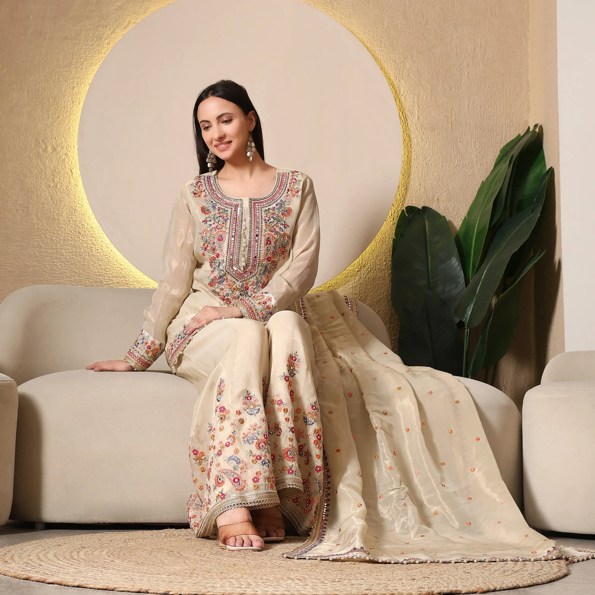 Woman in embroidered beige Anarkali suit with dupatta, sitting on sofa in elegant setting