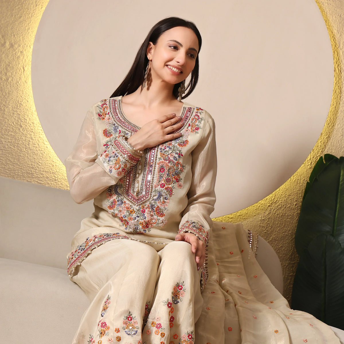 Woman wearing cream embroidered ethnic suit with floral patterns, sitting indoors, smiling.