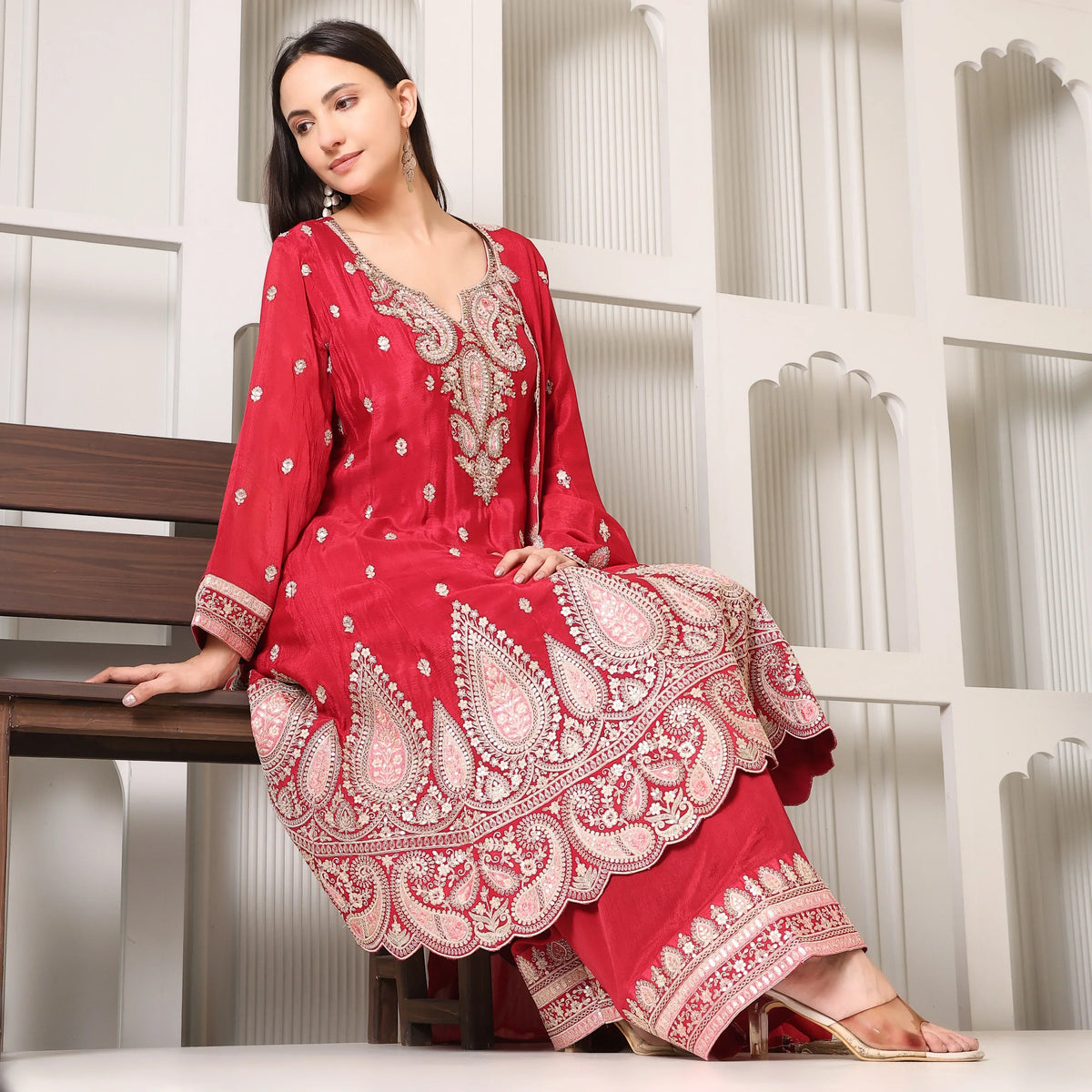 Woman in red Banarasi Anarkali suit with intricate embroidery, seated indoors
