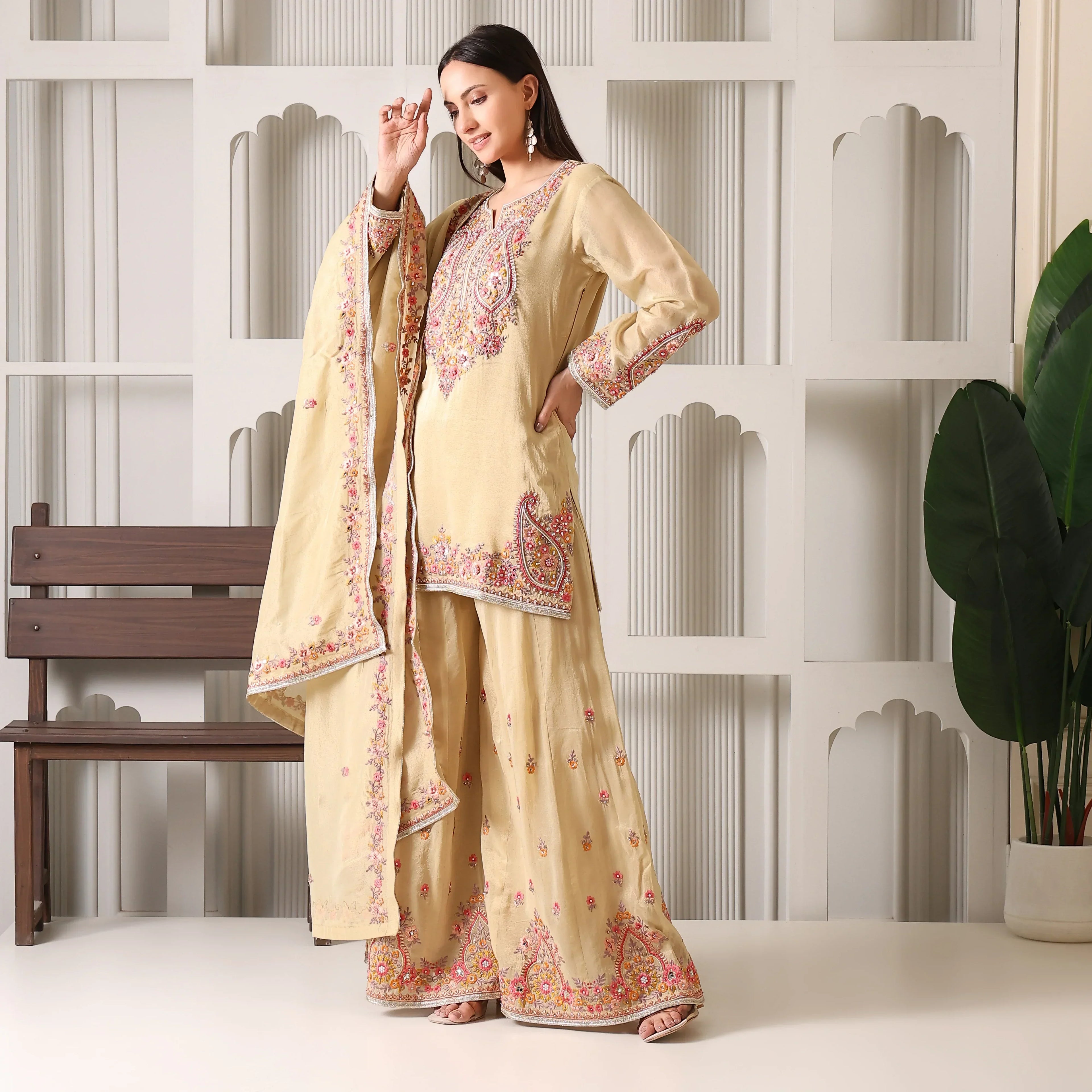 Woman wearing beige embroidered ethnic suit with dupatta, standing in a modern interior.