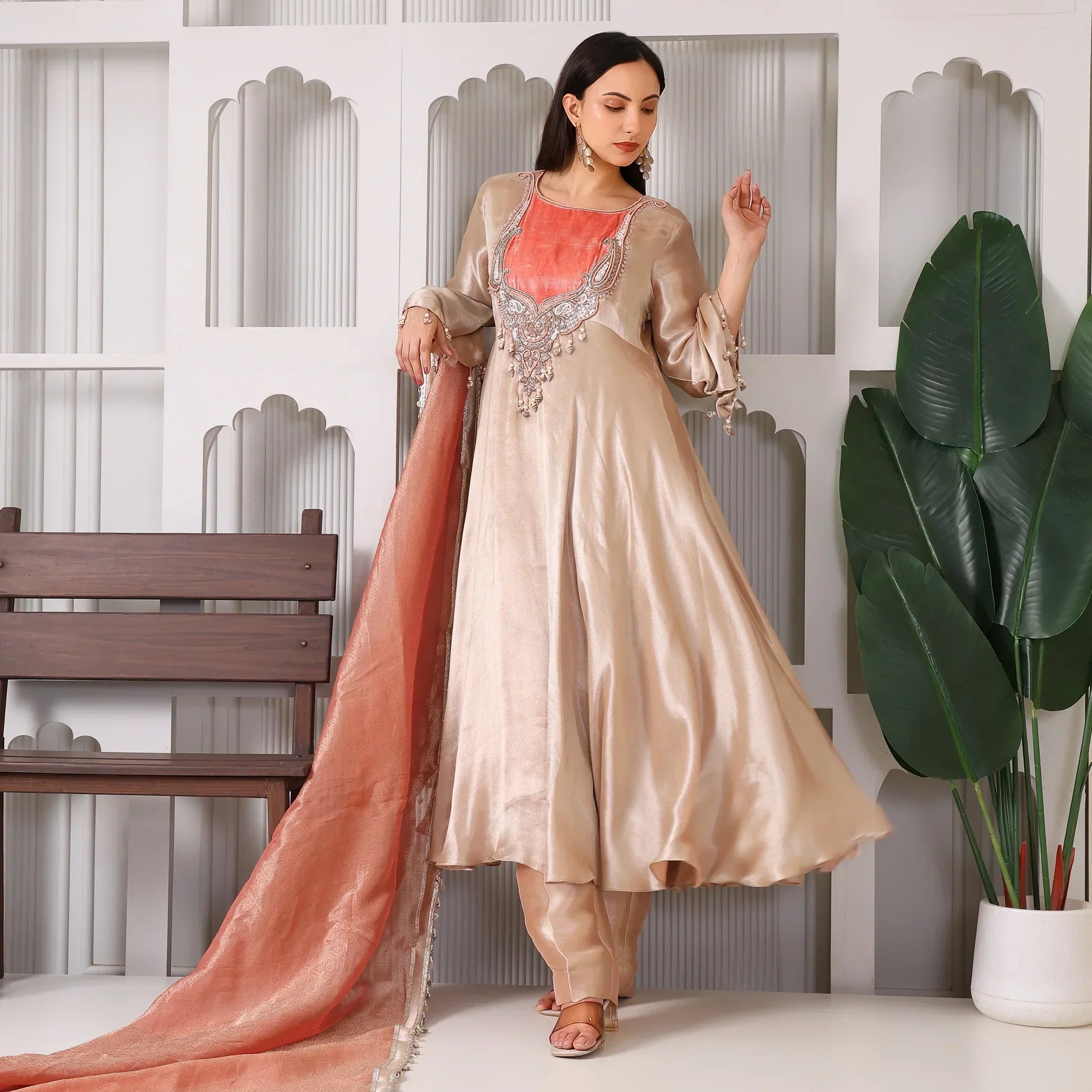 Woman in a gold silk Anarkali suit with peach dupatta, standing indoors beside plants.
