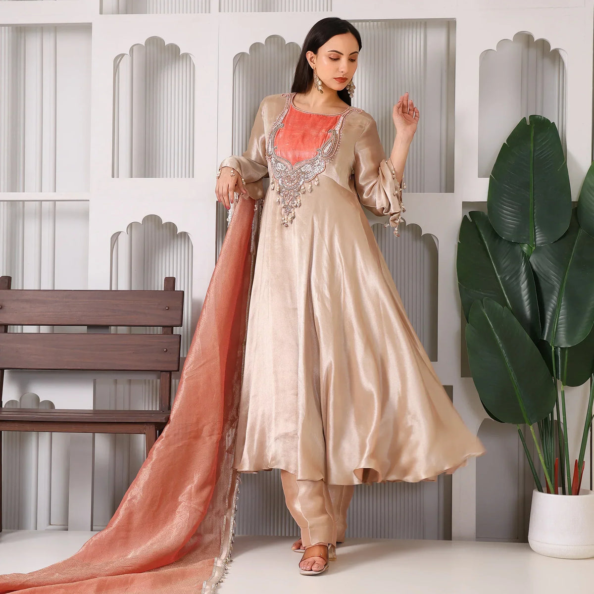 Woman in a gold silk Anarkali suit with peach dupatta, standing indoors beside plants.