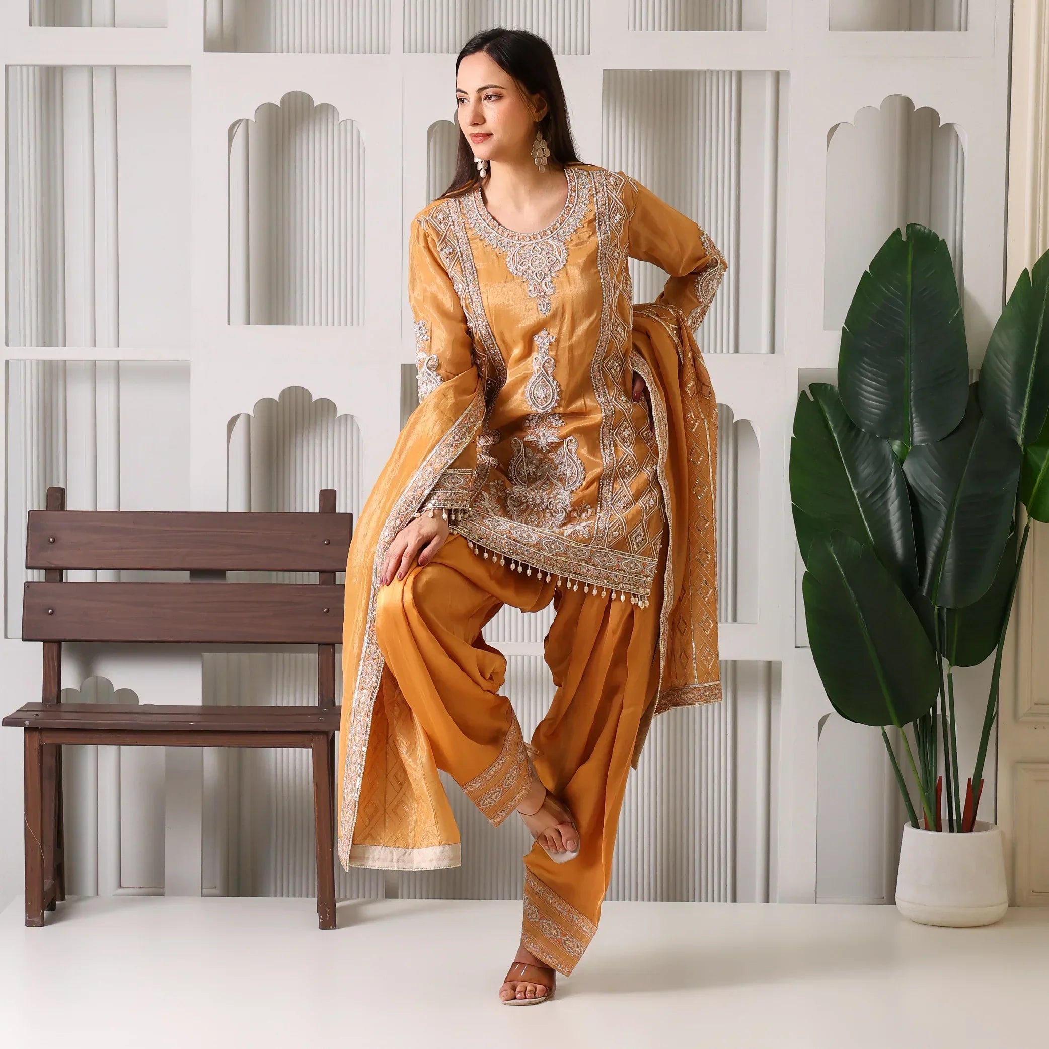 Woman in mustard yellow embroidered ethnic suit with dupatta, posing indoors near a bench and potted plant