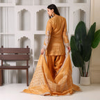 Woman in golden ethnic suit with embroidered dupatta, standing indoors by decorative wall.