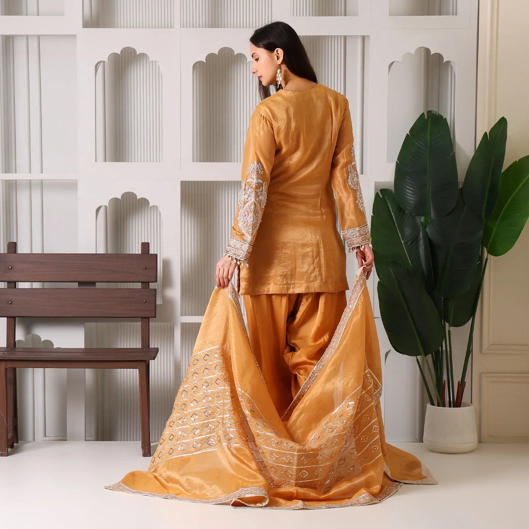Woman in golden ethnic suit with embroidered dupatta, standing indoors by decorative wall.