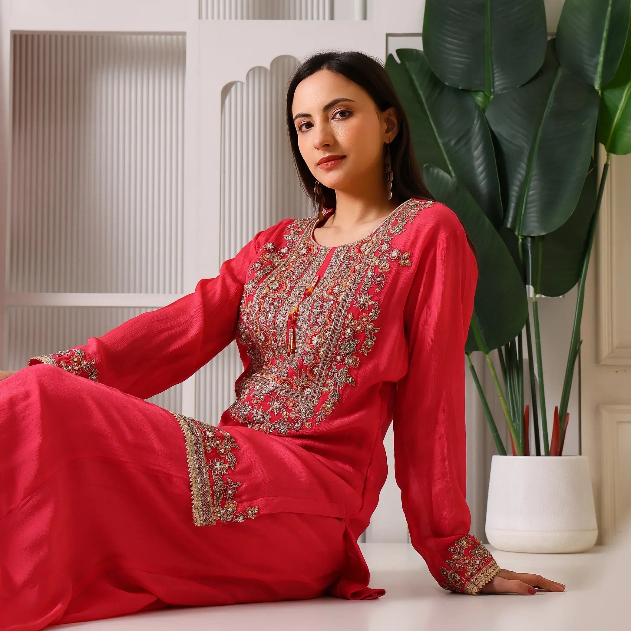 Woman in pink embroidered ethnic suit with gold accents, sitting indoors near green plant