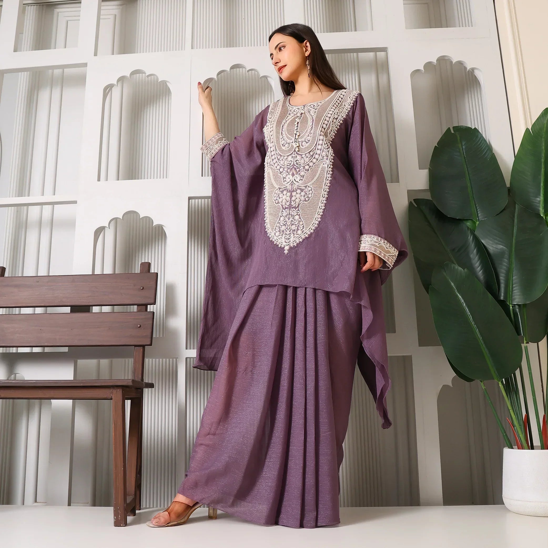 Woman in purple embroidered ethnic suit posing indoors with green plant and wooden bench