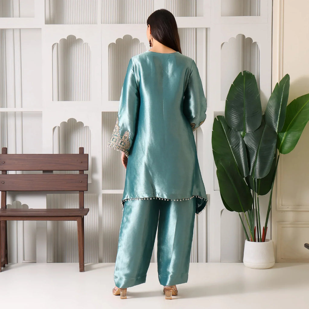 Woman in teal silk ethnic suit with embroidered sleeves, standing indoors near bench and plant