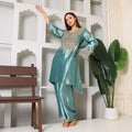 Woman in teal silk ethnic suit with intricate embroidery, posing indoors near a bench and plant.
