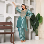 Woman in teal silk ethnic suit with intricate embroidery, posing indoors near a bench and plant.