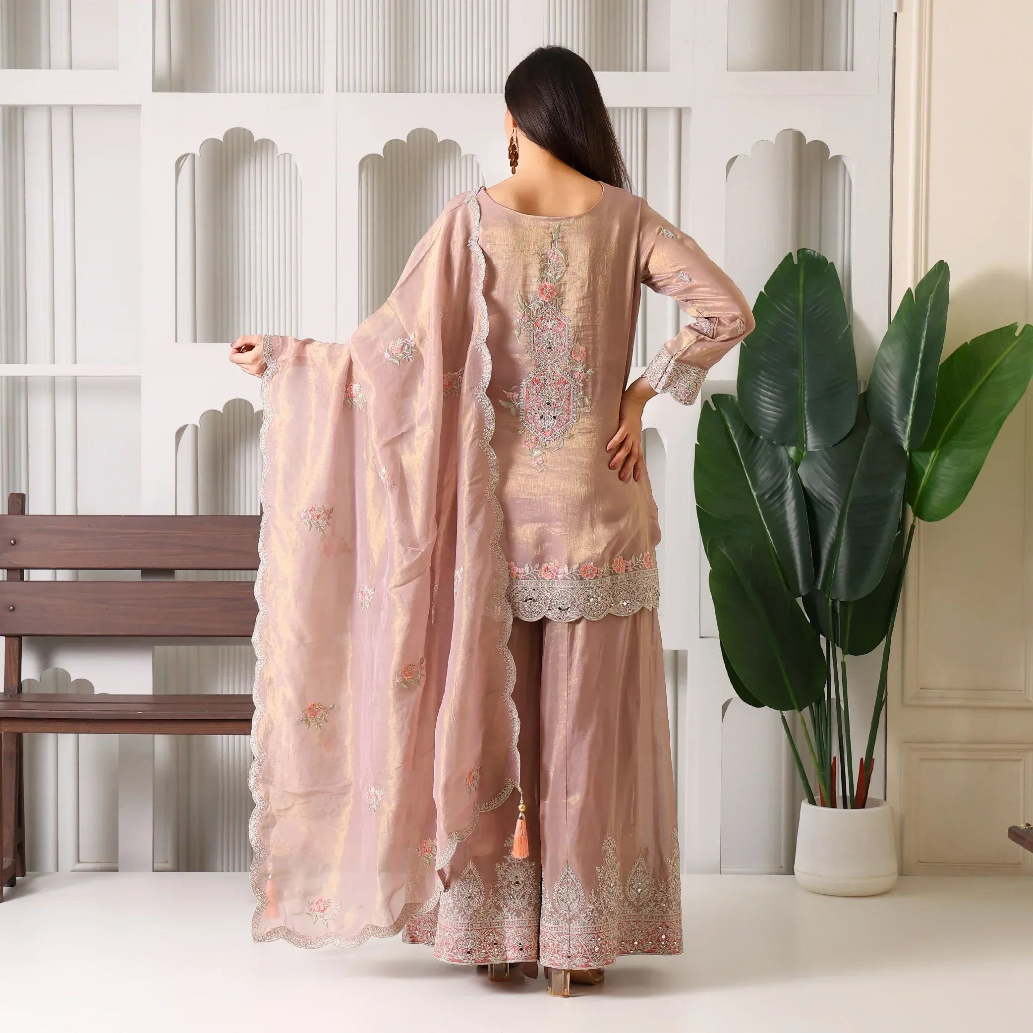 Woman in pastel pink embroidered ethnic sharara suit and dupatta, indoor setting with plants