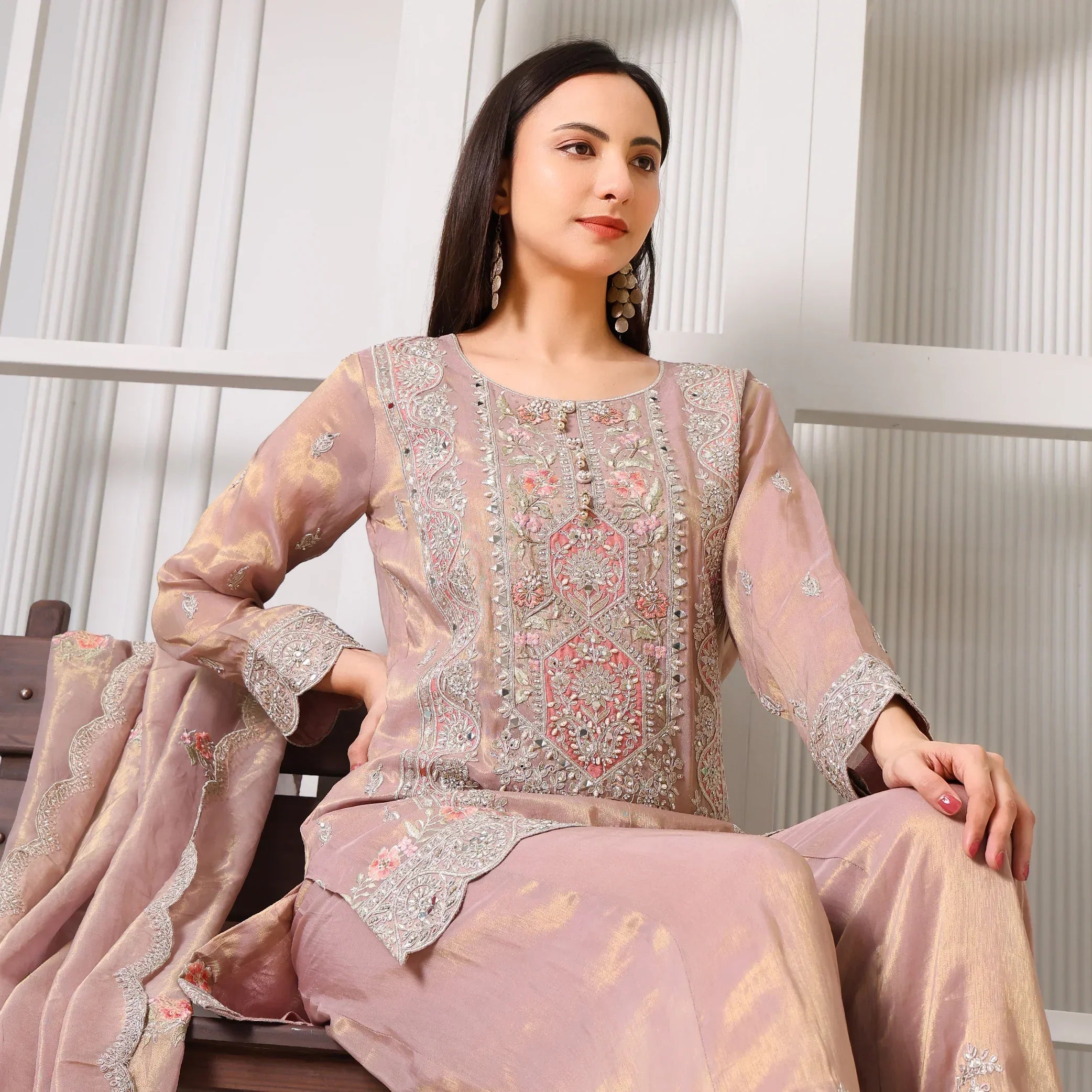 Woman wearing mauve embroidered ethnic suit with dupatta, sitting on bench indoors
