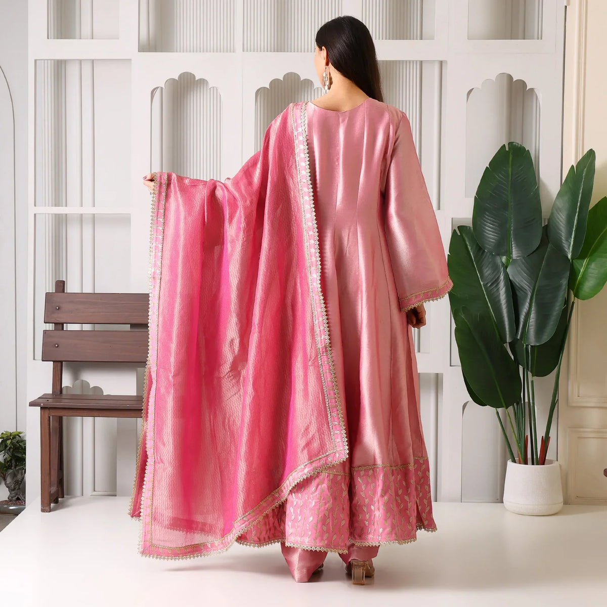 Woman in pink Banarasi Anarkali suit with dupatta, standing indoors by green plant