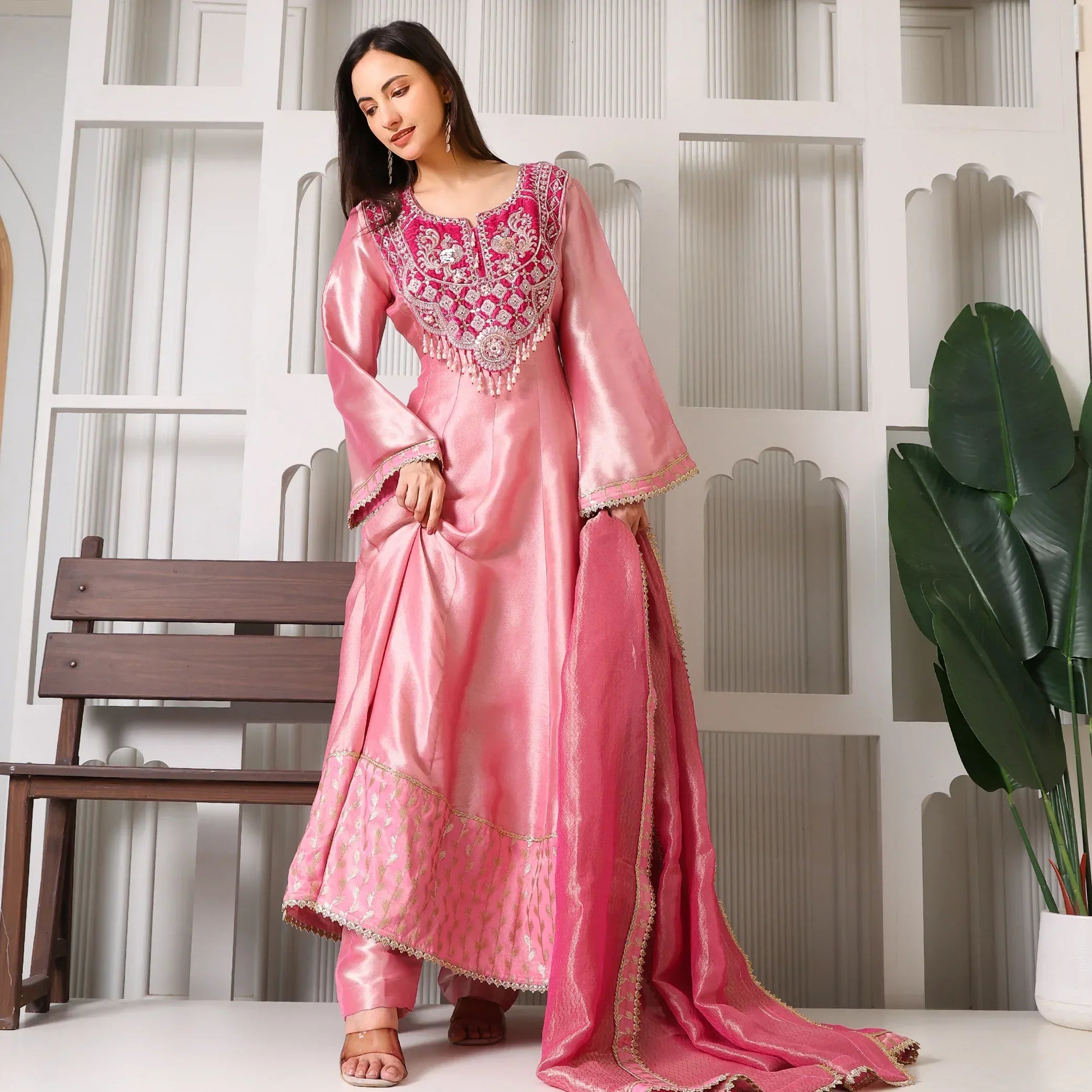 Woman wearing a pink Banarasi Anarkali suit set with embroidery, posed indoors by a bench