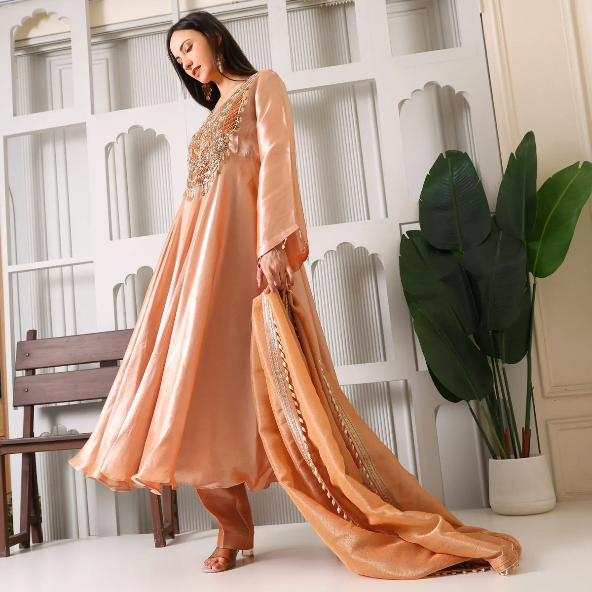 Woman in peach Banarasi Anarkali suit with dupatta, standing indoors near plants