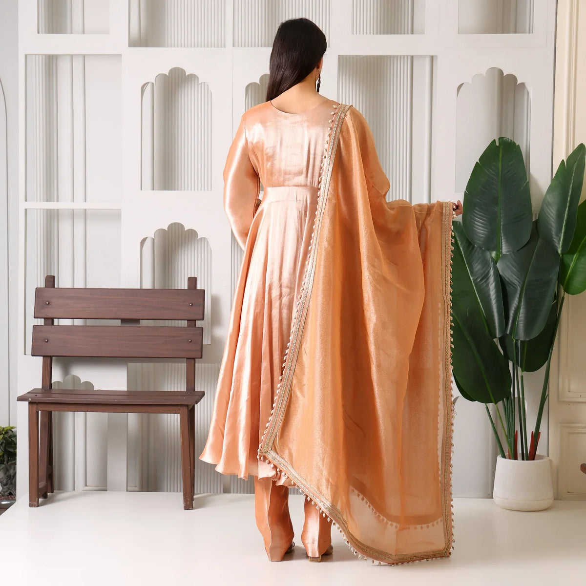 Woman in peach Banarasi Anarkali suit with dupatta, standing indoors by a bench and plant