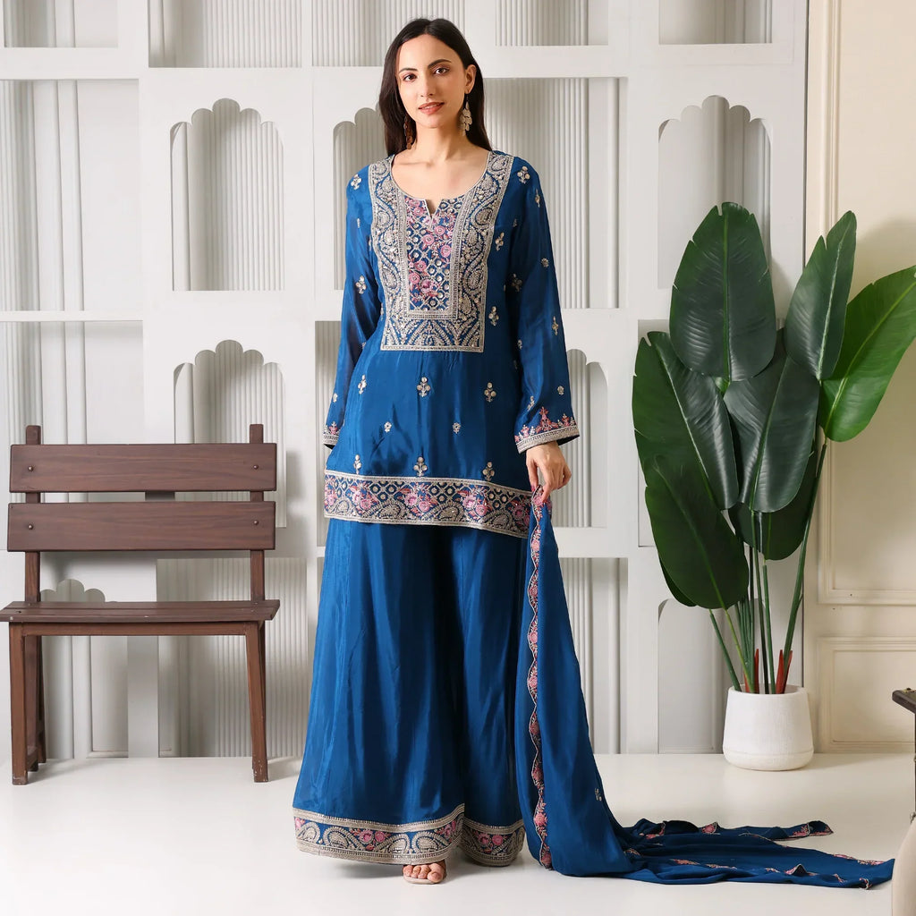 Woman in a blue embroidered ethnic suit set with dupatta, standing indoors near a plant.