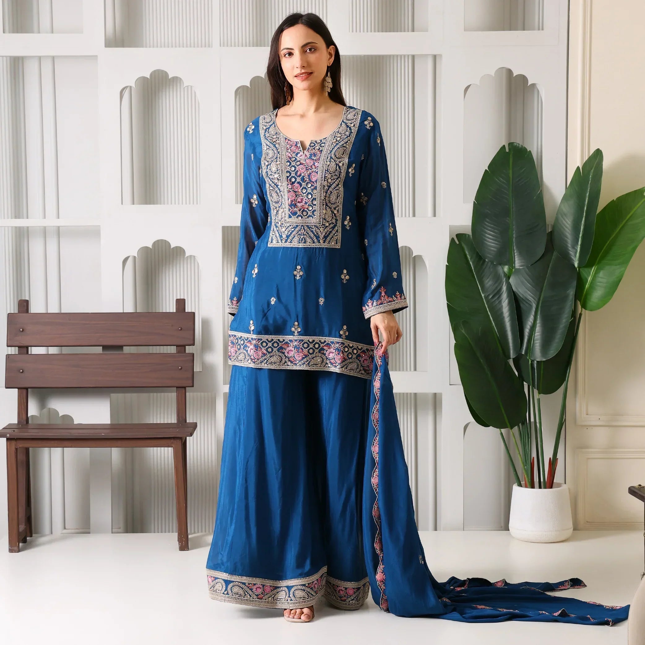 Woman in a blue embroidered ethnic suit set with dupatta, standing indoors near a plant.