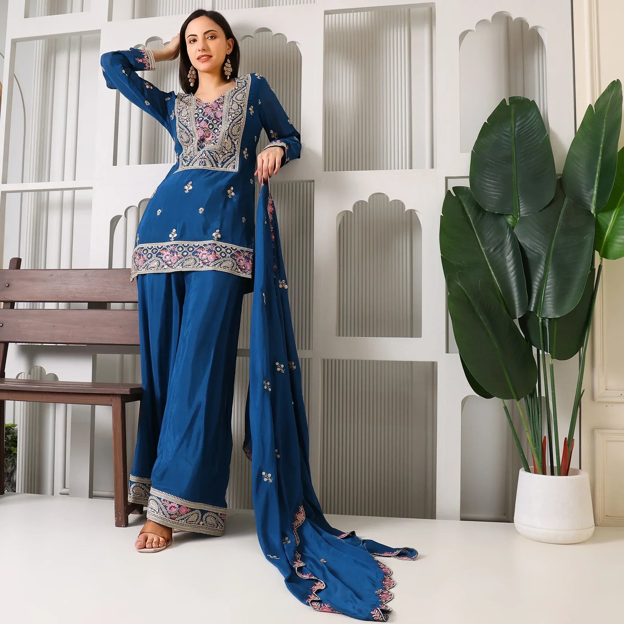 Woman in blue embroidered ethnic suit with dupatta, standing indoors by white wall and plant