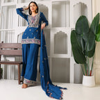Woman in blue embroidered ethnic suit with dupatta, standing indoors by white wall and plant