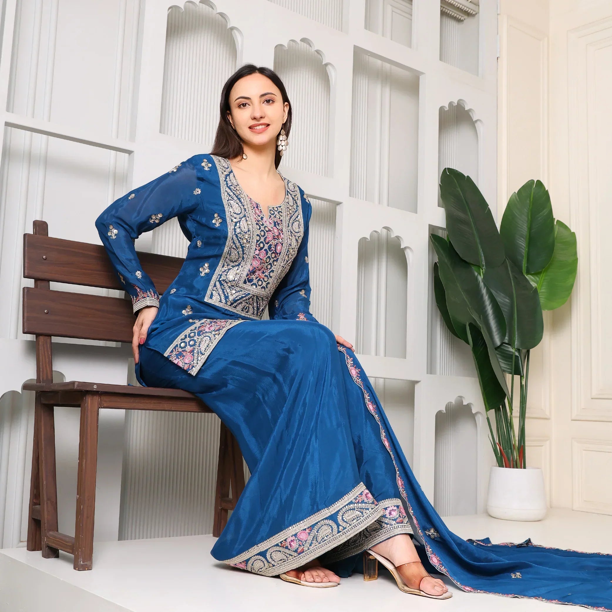 Woman in blue embroidered Anarkali suit with dupatta, sitting on a bench, indoor setting
