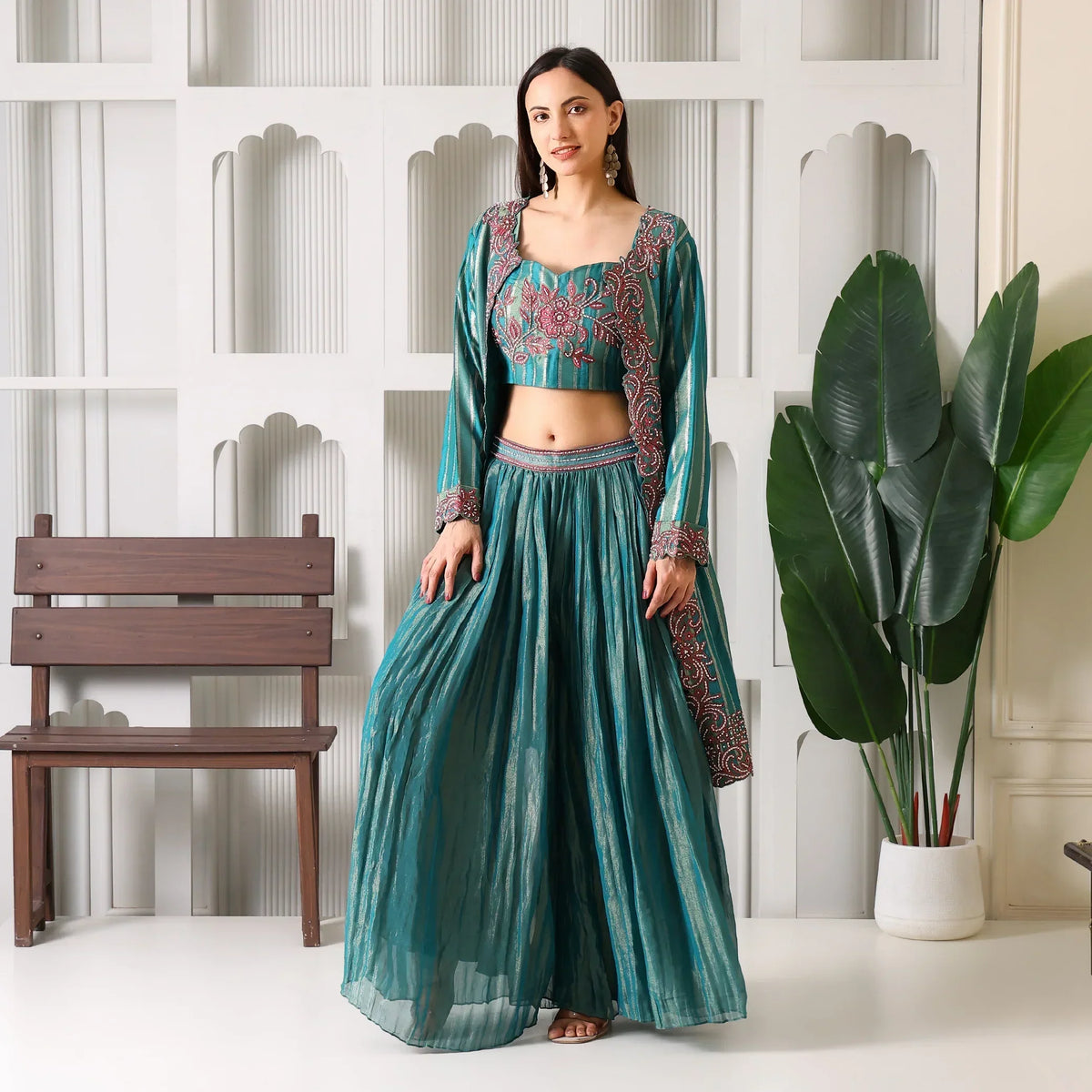 Woman in teal embroidered ethnic suit set with flared pants, standing indoors by a plant