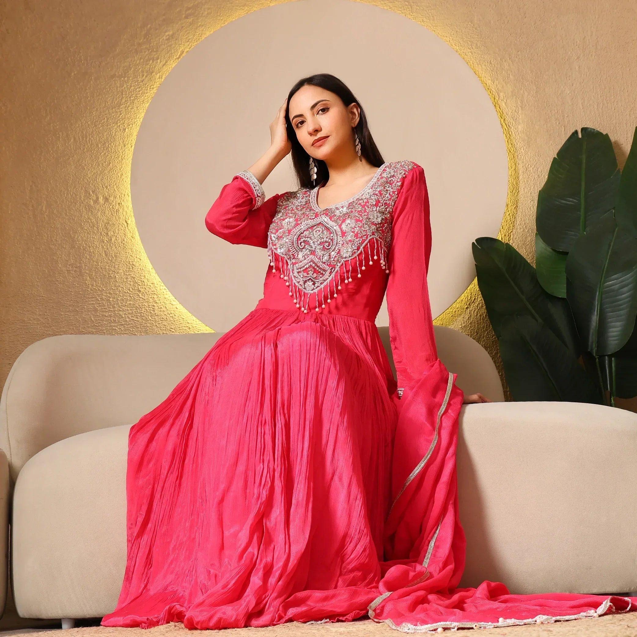 Woman in a bright pink embroidered Anarkali suit seated on a beige sofa with golden backdrop