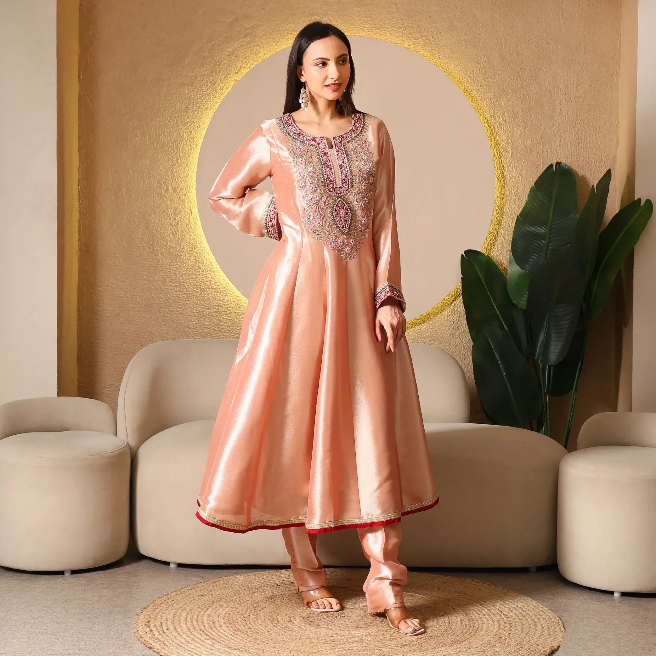 Woman wearing a peach Banarasi Anarkali suit with intricate embroidery, standing in a modern living room.