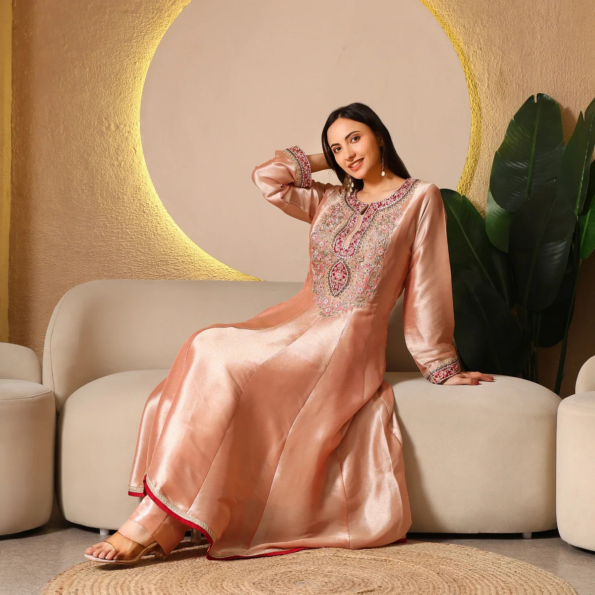 Woman in peach silk Anarkali suit with intricate embroidery, sitting on a sofa