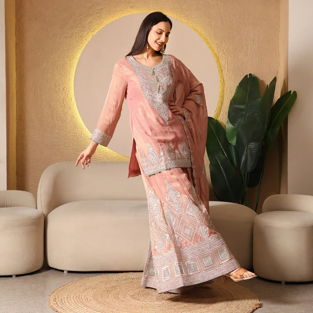 Woman in pastel pink embroidered Anarkali suit with dupatta, posing in modern living room