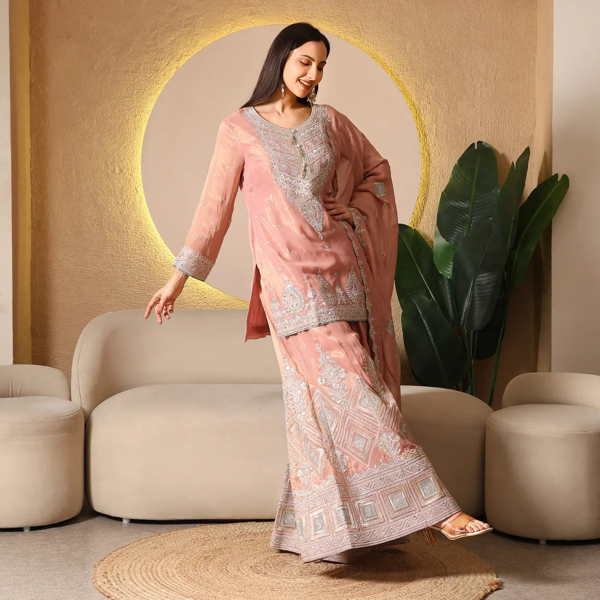 Woman in pastel pink embroidered Anarkali suit with dupatta, posing in modern living room