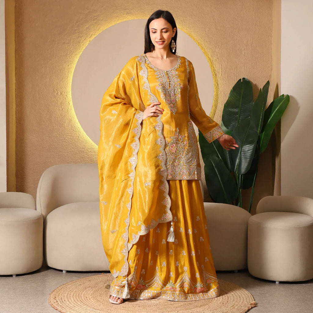 Woman in mustard yellow embroidered ethnic suit with dupatta, standing in modern living room