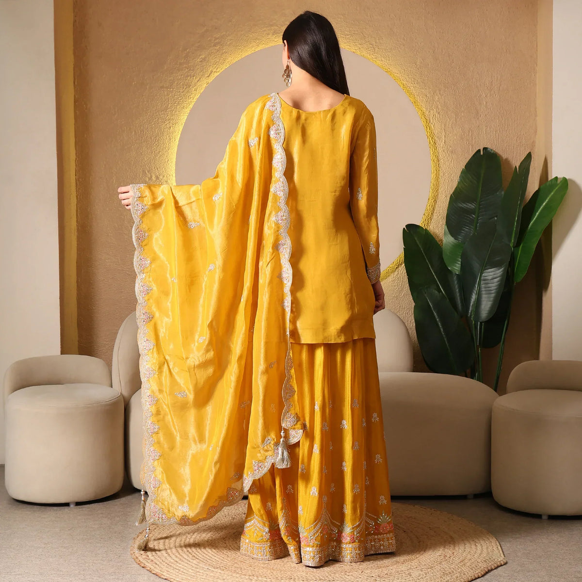 Woman in mustard ethnic suit with embroidered dupatta and sharara, standing indoors