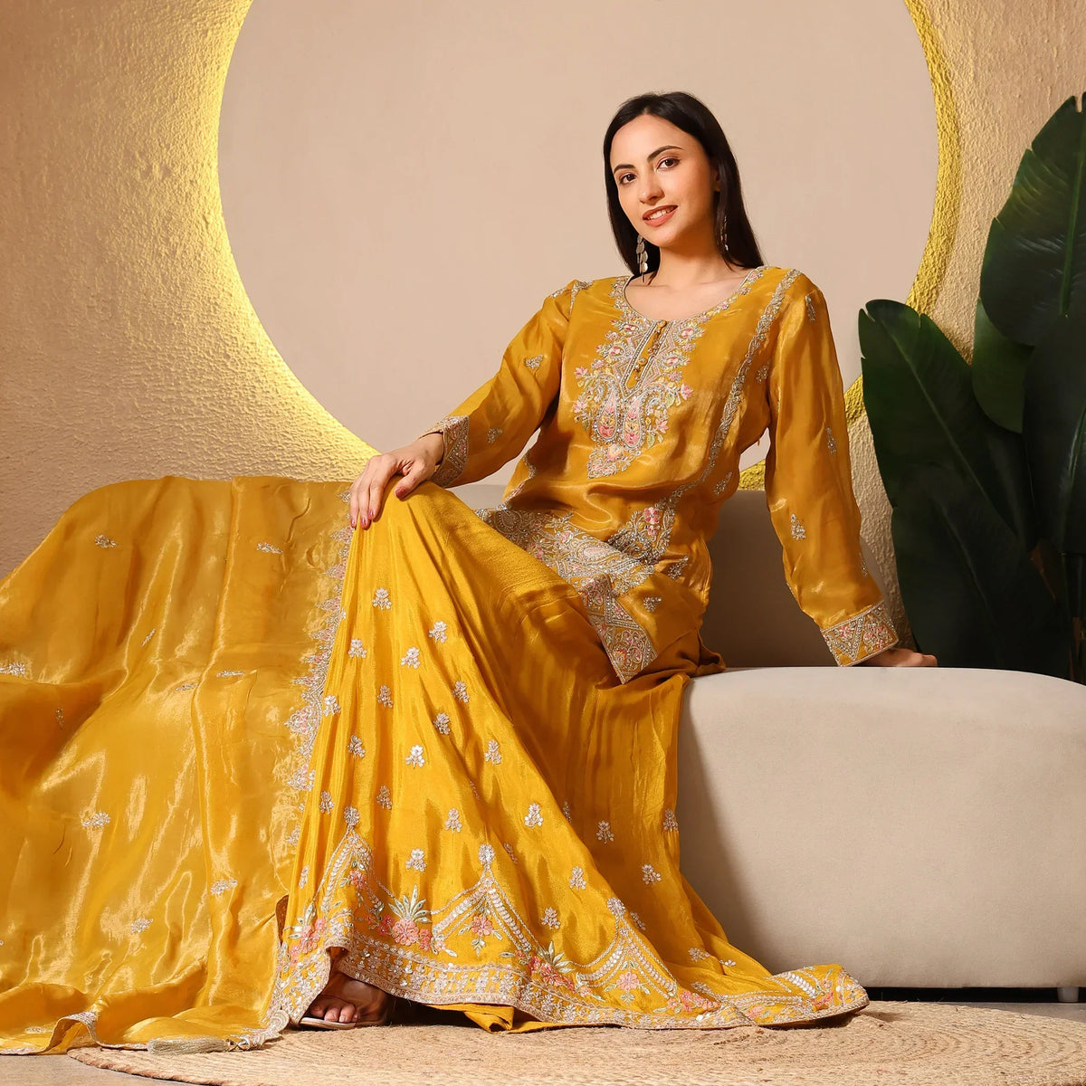 Woman wearing yellow embroidered ethnic suit with dupatta, sitting on beige sofa