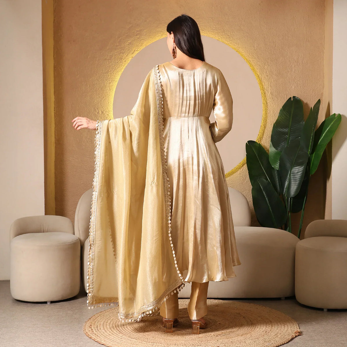 Woman in gold Banarasi Anarkali suit with dupatta, standing in elegant modern room