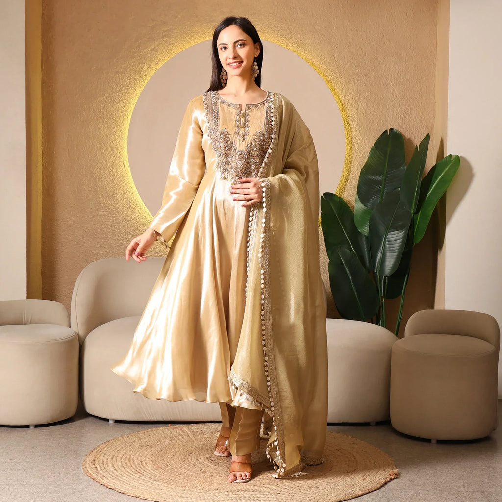 Woman in gold Banarasi Anarkali suit with dupatta, standing in a modern living room